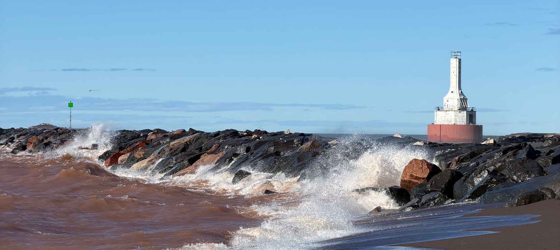 Waves breaking over a rocky Lake Superior breakwater, with a lighthouse standing at its far end.