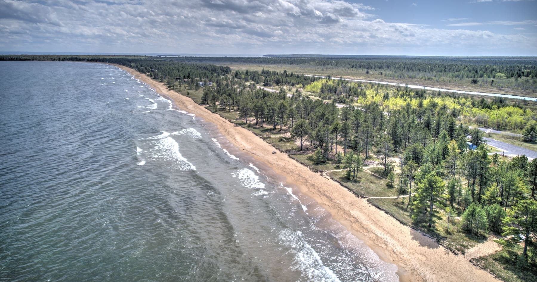 Aerial view of a long Lake Superior shoreline, waves breaking on sand beside a pine forest.
