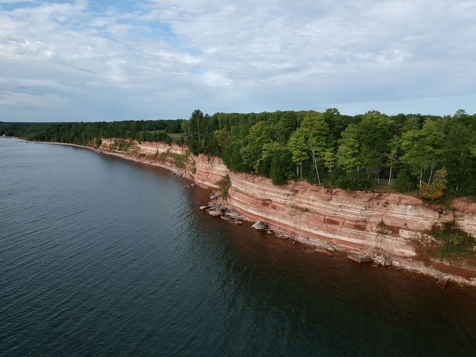 Aerial view of layered red sandstone cliffs and forested shoreline meeting the deep water of Lake Superior.